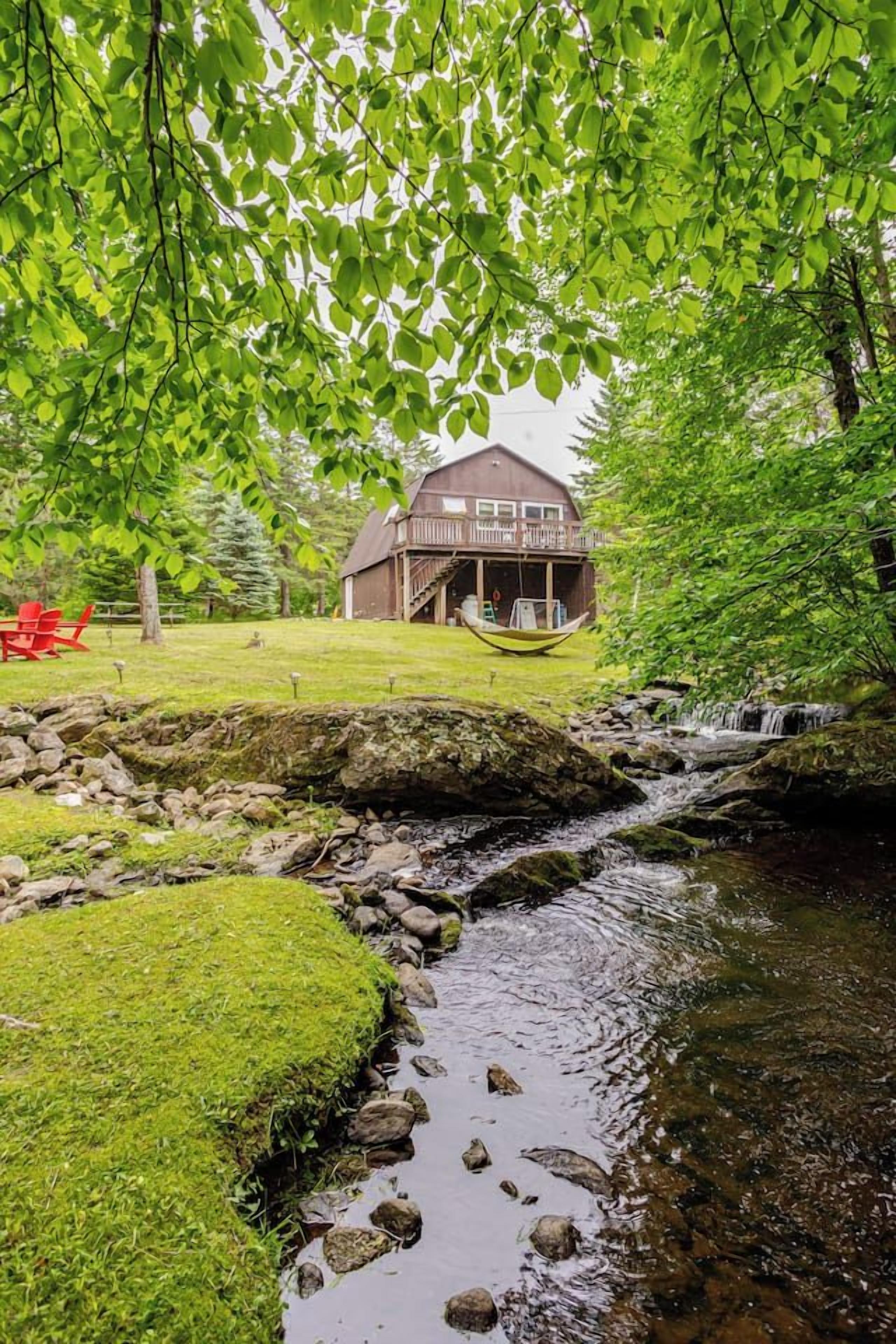 A rustic cabin with a porch, a small stream, and a grassy area with red chairs.