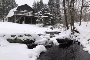A cabin with a deck, surrounded by snow-covered trees and a stream.