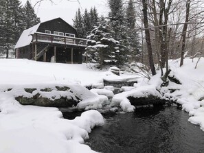 A cabin with a deck and a small stream flowing through a snowy landscape.
