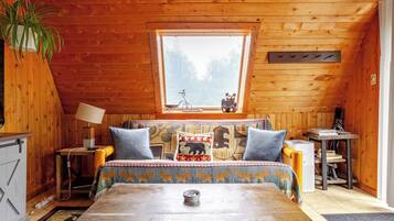 A cozy cabin interior with a wooden ceiling, a bed with patterned bedding, a small table, and a window with a view of trees.