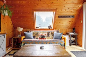 Ein gemütliches Blockhausschlafzimmer mit Holzbalkendecke, einem Bett mit gemusterten Textilien, einem kleinen Tisch und einem Fenster mit Blick auf Bäume.