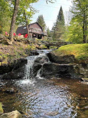 Ein rustikales Holzhäuschen mit einer Holzterrasse und einem kleinen Wasserfall im Vordergrund.