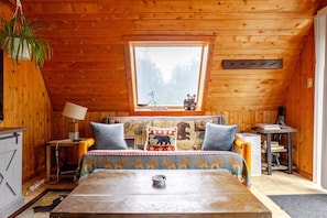A cozy cabin room with a wooden ceiling, a large window, a bed with a patterned bedspread, a table, and a chair.