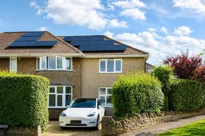A two-story house with solar panels on the roof, a white car parked in the driveway, and a well-maintained hedge.