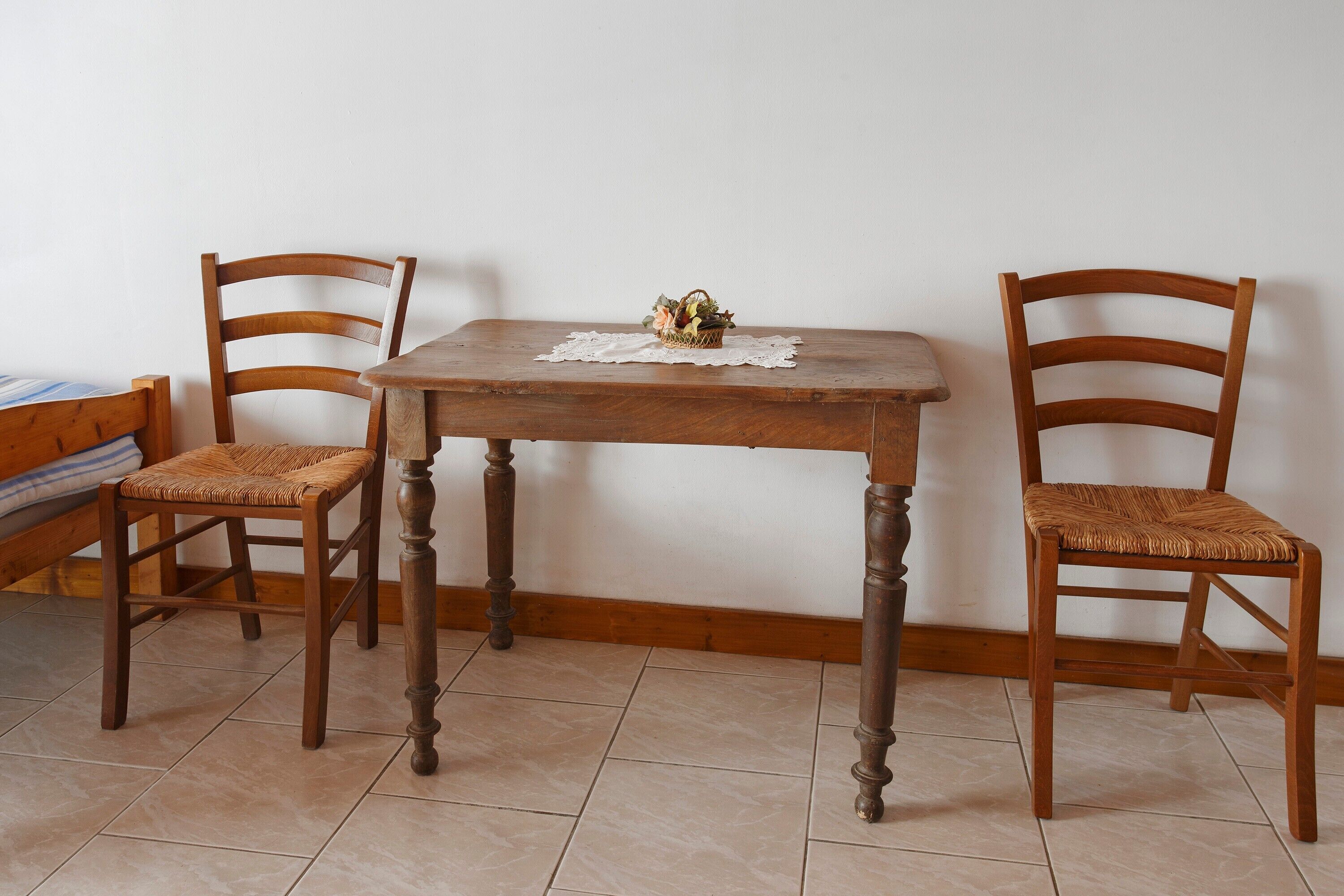 Une table de salle à manger en bois, accompagnée de deux chaises, d’une nappe en dentelle et d’un panier rempli de fleurs.