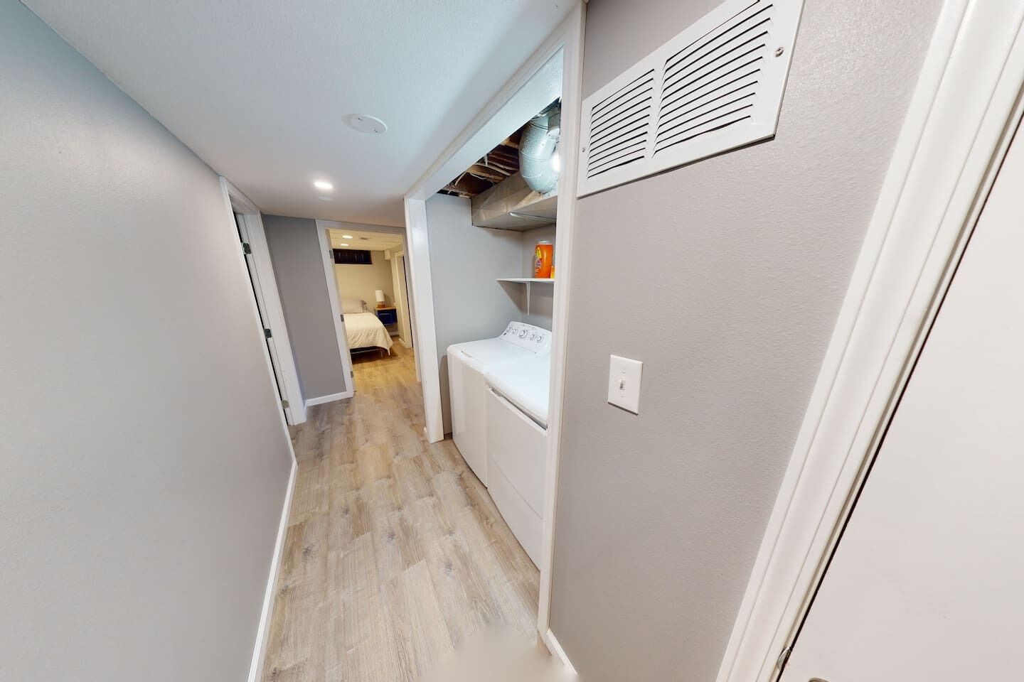 A narrow hallway with a laundry area on the right, a bed in the background, and a vent on the wall.