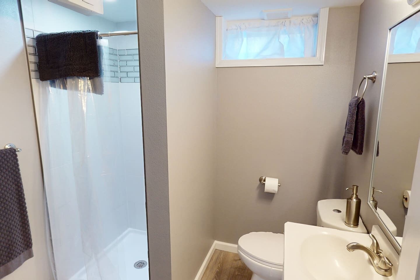 Une salle de bain avec une douche à parois de verre, des toilettes, un lavabo avec un robinet doré et une fenêtre ornée de rideaux légers.