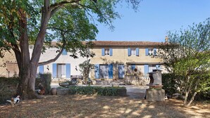 A two-story building with a tiled roof, blue shutters, and a garden with a large tree and a bird bath. - Guest Room "Les Lilas" with Shared Terrace, Communal Garden and Wi-Fi (Antezant-la-Chapelle)
