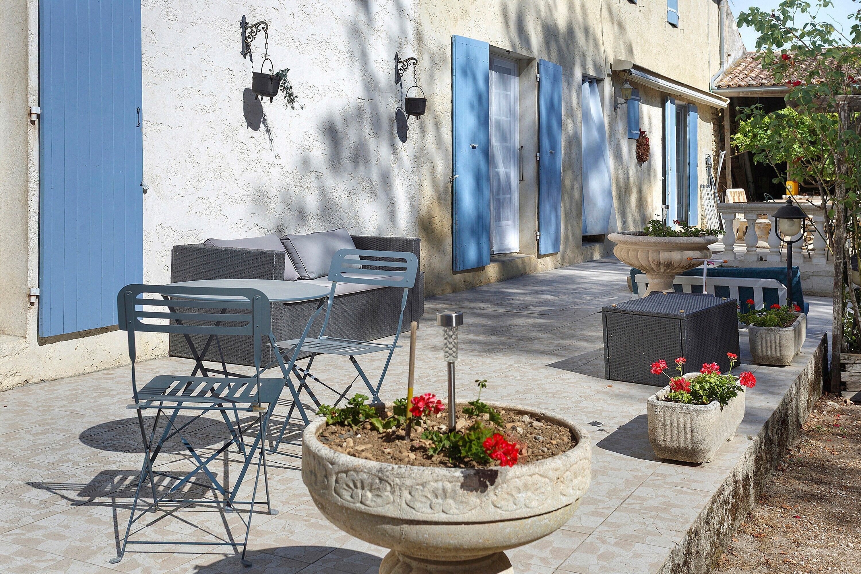 Un espace extérieur aménagé avec des tables et des chaises, un grand bac à fleurs et un bâtiment aux volets bleus.