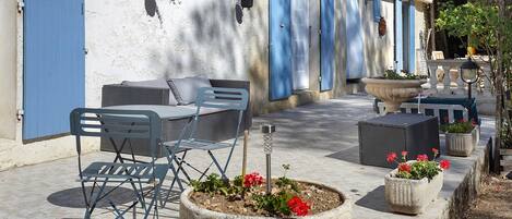An outdoor seating area with tables and chairs, a large planter with flowers, and a building with blue shutters.