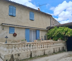 A two-story building with blue shutters, a balcony with a balustrade, and a garden with potted plants.