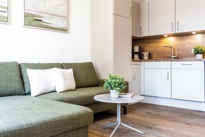 Ein moderner Wohnraum mit einem grünen Sofa, einem kleinen runden Tisch mit Pflanze und einer Küchenzeile mit weißen Schränken und einer Holz Arbeitsplatte.