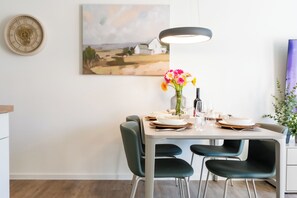 Een eethoek met een tafel voor vier personen, stoelen, een bloevas, een fles wijn en een schilderij aan de muur.