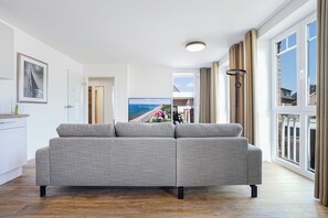 A modern living room with a grey sofa, a flat-screen TV, and a view of a beach through large windows. - Dünenanker 12. Infrared cabin. Roof terrace. Central location. (Grömitz)