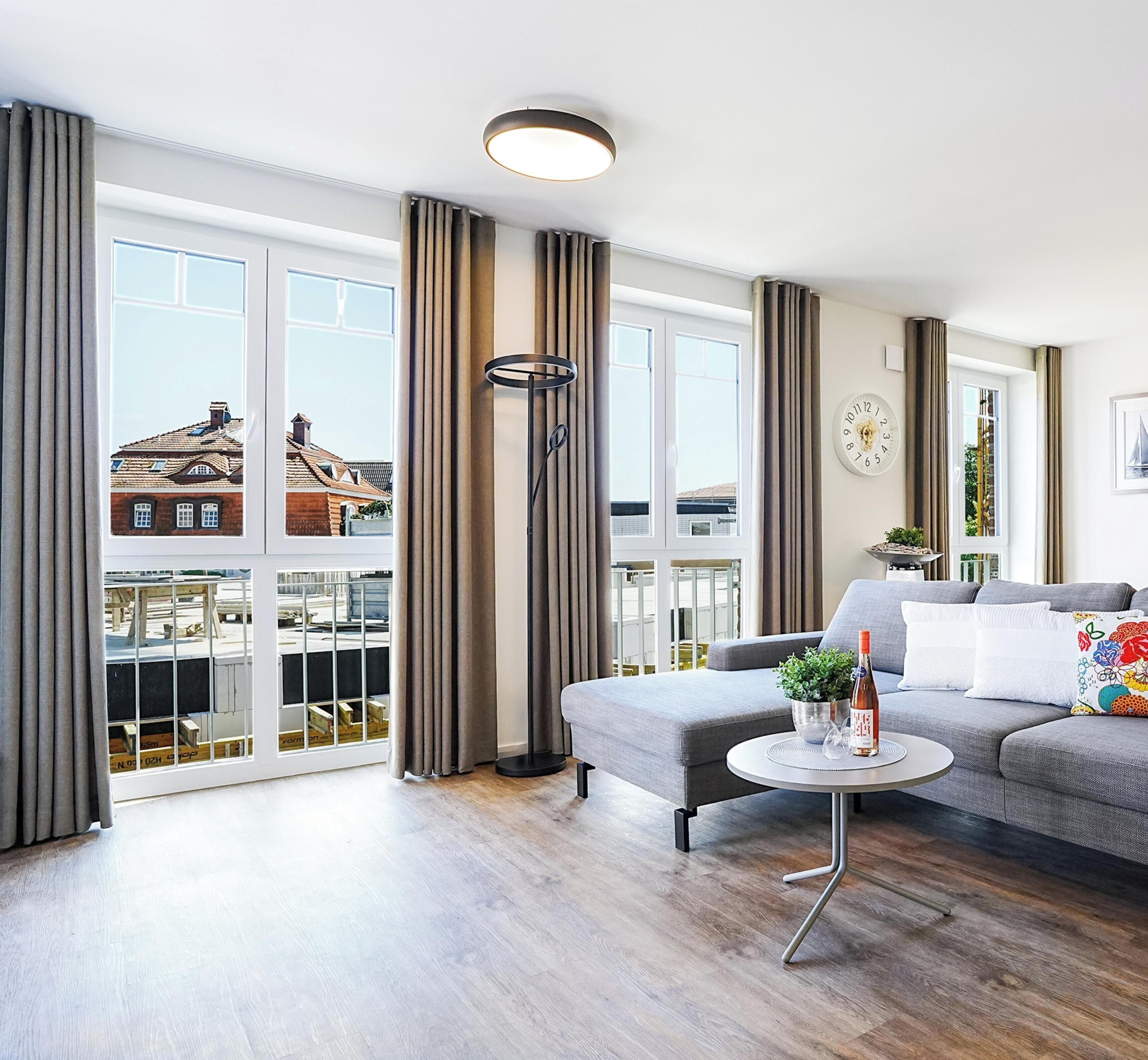 Un salon moderne avec un canapé gris, une table basse ronde et une vue sur les immeubles à travers de grandes fenêtres.