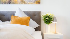 En snyggt bäddad säng med ett vitt täcke och en senapsgul kudde, en grå klädd sänggavel och ett nattduksbord med en vit vas med blommor och en lampa.