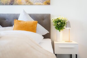Ein ordentlich bezogenes Bett mit weißer Decke und einem senfgelben Kissen, ein grauer gepolsterter Kopfteil und ein Nachttisch mit weißer Blumenvase und einer Lampe.