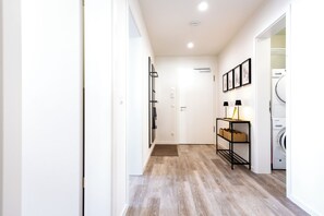 Ein Flur mit Holzboden, einer Tür, einem Spiegel, einem an der Wand befestigten Regal mit Körben, einer Lampe und einer Waschmaschine.