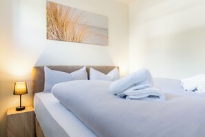 Ein ordentlich bezogenes Bett mit weißen Leinentüchern und einem gefalteten Handtuch, eine Nachttischlampe und ein Gemälde mit hohem Gras an der Wand.