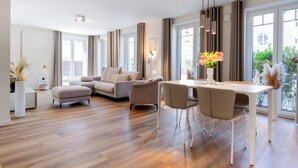 A modern living room with a sofa, a dining table with chairs, and a view of the outdoors through large windows. - MeerNah Grömitz - Apartment 1. terrace. Sauna. Family friendly. (Grömitz)