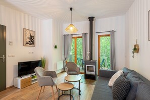 Ein modernes Wohnzimmer mit Flachbildfernseher, einer Couch, Sesseln, einem kleinen Tisch, einem Kamin und einem großen Fenster mit Vorhängen.