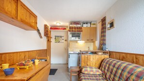 Eine kompakte Küche mit Holdekor-Schränken, einem kleinen Kühlschrank und einer Mikrowelle. Ein Esstisch mit einer Bank und ein farbenfrohes Karomuster-Sofa sind ebenfalls vorhanden.