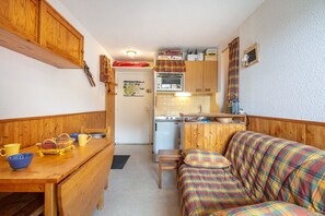 A compact kitchen with wooden cabinets, a small refrigerator, and a microwave. There is a dining table with a bench and a colorful plaid sofa. - CosyPeak Alpe d’Huez - Au pied des pistes (Huez)
