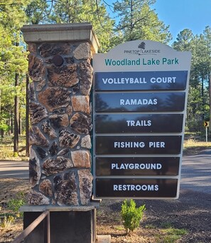 Señal que enumera las comodidades del Parque Woodland Lake, incluyendo cancha de voleibol, pérgolas, senderos, muelle de pesca, parque infantil y baños.