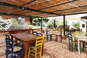 Um pátio ao ar livre com mesas e cadeiras de madeira, rodeado por vasos de plantas e flores.