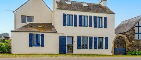 Un bâtiment à deux étages, doté d’une façade blanche et de volets bleus, avec une entrée en arc de pierre et un toit en pente.