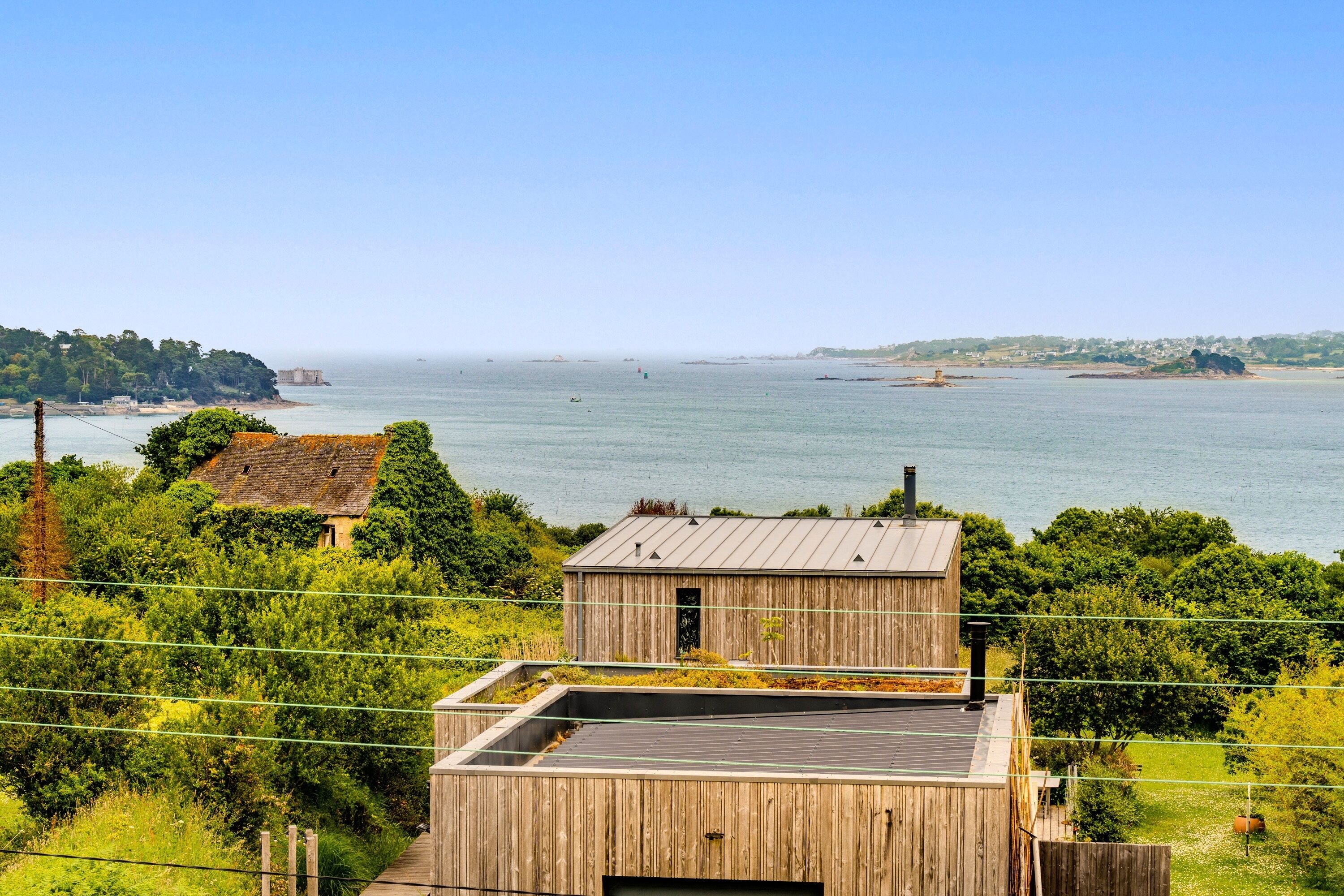 A wooden building with a flat roof, surrounded by greenery and overlooking a large body of water.