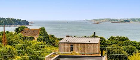 A wooden building with a flat roof, surrounded by greenery and overlooking a large body of water.