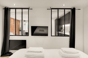 Ein modernes Hotelzimmer mit einem großen Bett, zwei schwarzen Vorhängen, einem Flachbildfernseher und Blick auf eine Ziegelwand sowie ein weiteres Zimmer.