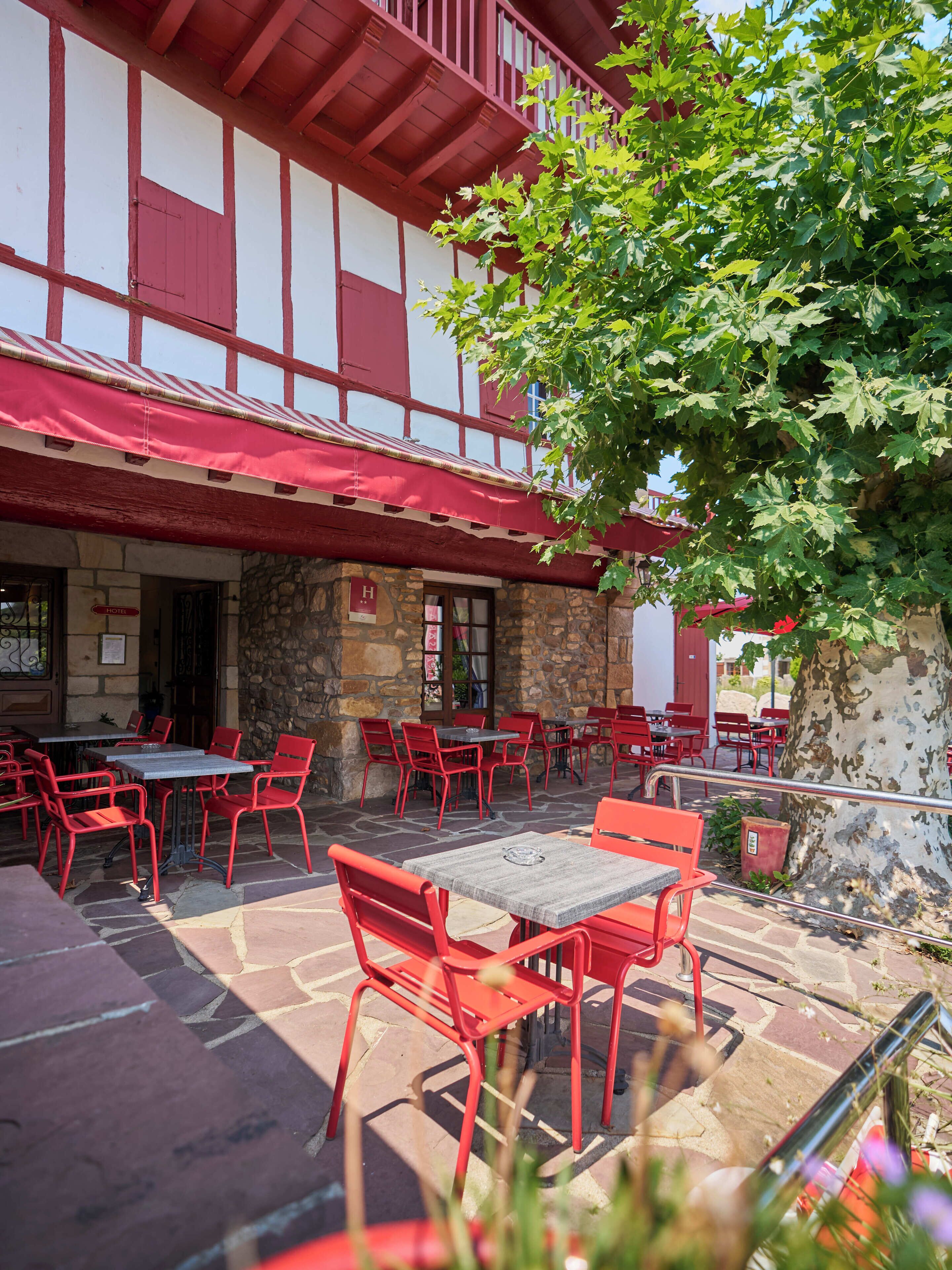 Un edificio de piedra con fachada roja y blanca, mesas y sillas rojas en la terraza, y un árbol en primer plano.