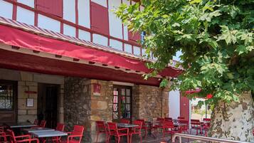 Un edificio de piedra con fachada roja y blanca, mesas y sillas rojas en la terraza, y un árbol en primer plano.