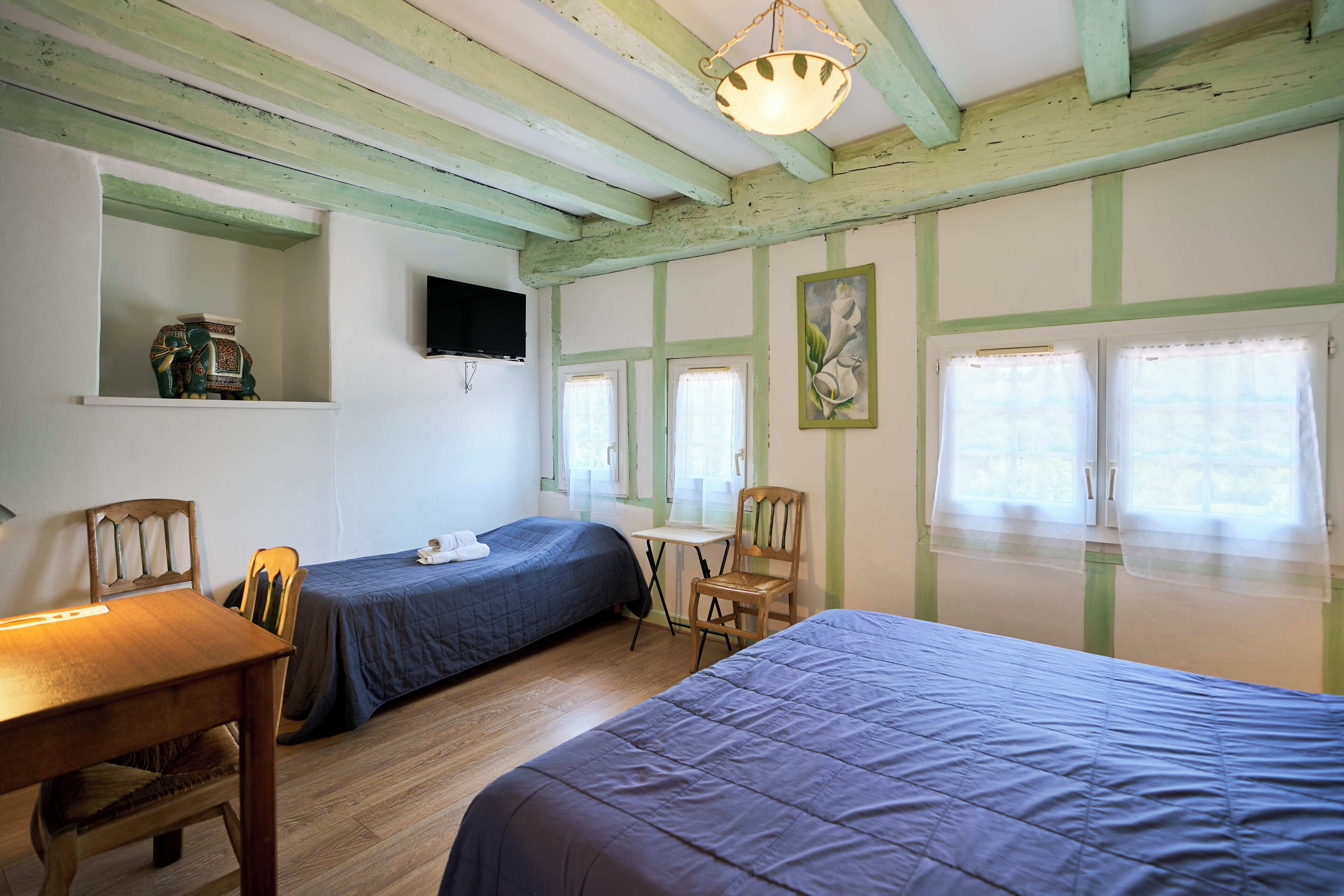 A room with two beds, a wooden table, a chair, a small desk, a television, and a window with curtains.
