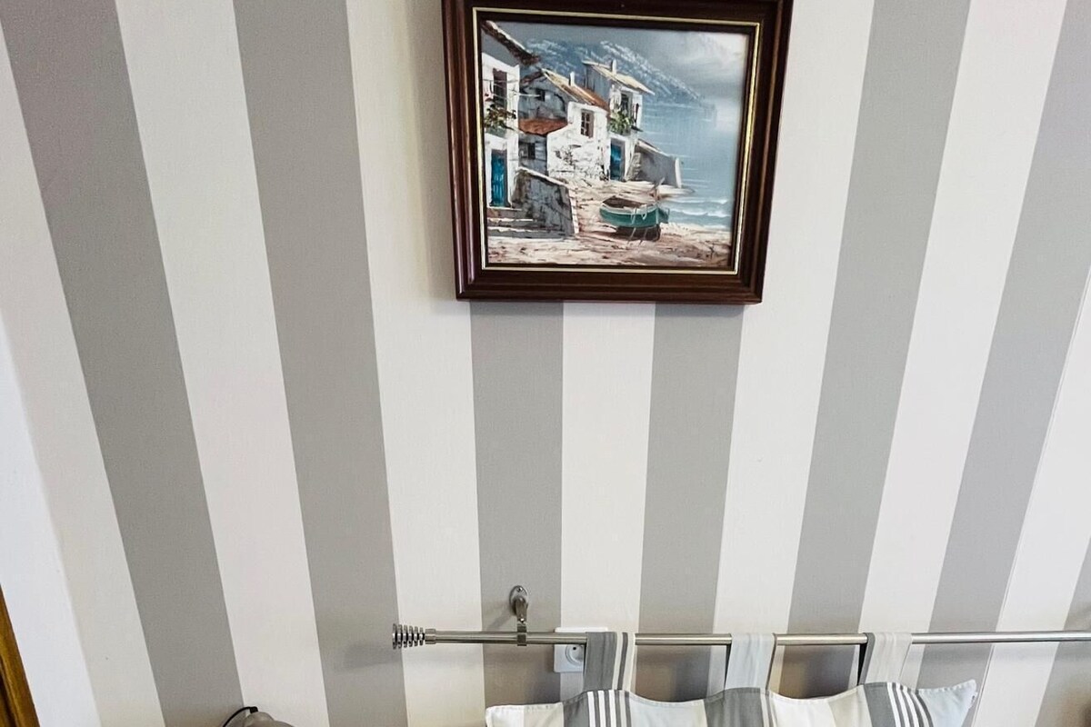 Una habitación con cama, mesita de noche con lámpara, pared a rayas y un cuadro enmarcado sobre la cama.