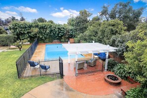 Ein Garten mit Schwimmbad, einer überdachten Terrasse und einer Feuerstelle aus Ziegeln.