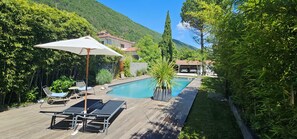 A pool area with lounge chairs and an umbrella, surrounded by greenery and a mountain backdrop. - Villa Blanche - Entre montagne, piscine et calme (Sévrier)
