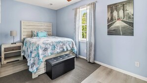 Ein Schlafzimmer mit Bett, Nachttisch, Deckenventilator, Fenster mit Vorhängen und einem Bild an der Wand.