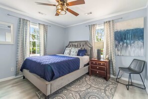 A bedroom with a bed, a nightstand, a chair, a ceiling fan, and a large abstract painting.