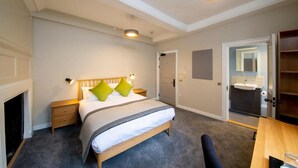 A bedroom with a bed, bedside tables, a nightstand, a desk, a chair, a fireplace, a door, and a bathroom with a sink. - Turl Street Mitre (Oxford)