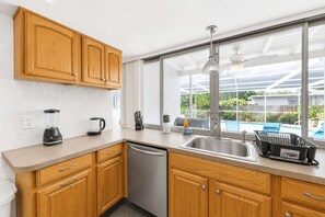 Une cuisine avec des meubles en bois, un lave-vaisselle, une machine à café et une fenêtre donnant sur une piscine.