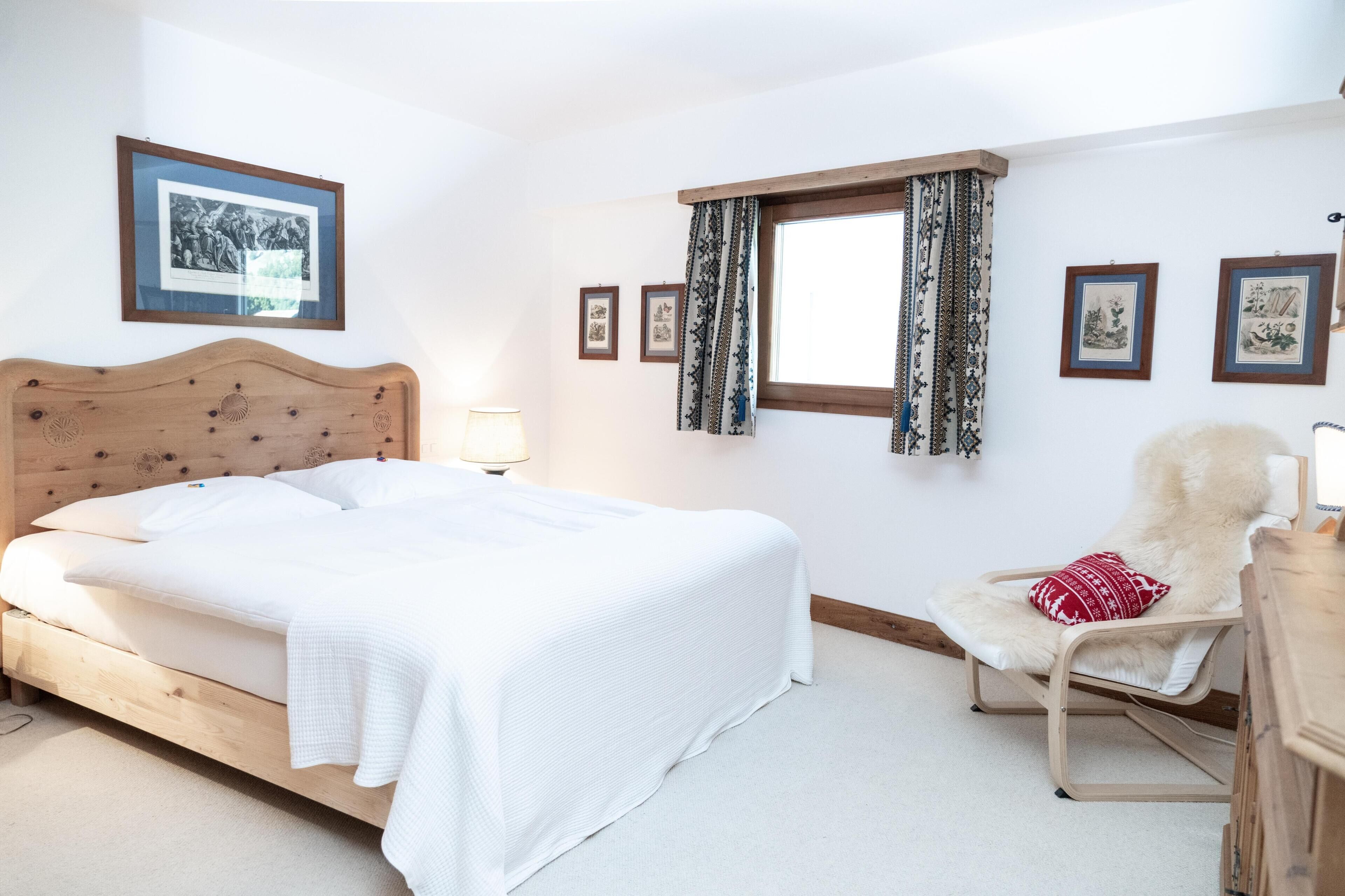 Een slaapkamer met een houten bed, een fauteuil met een bont plaid en ingelijste foto’s aan de muur.