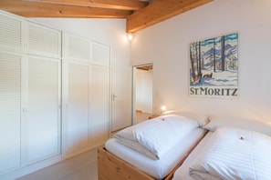 Ein Schlafzimmer mit Holzbalkendecke, weißen Betten, einem Kleiderschrank und einem Poster mit einer verschneiten Berglandschaft.