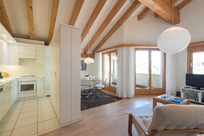 Eine moderne Küche mit weißen Schränken, ein Essbereich mit Tisch und Stühlen, ein Wohnzimmer mit Sofa und Fernseher sowie Holzbalken an der Decke.