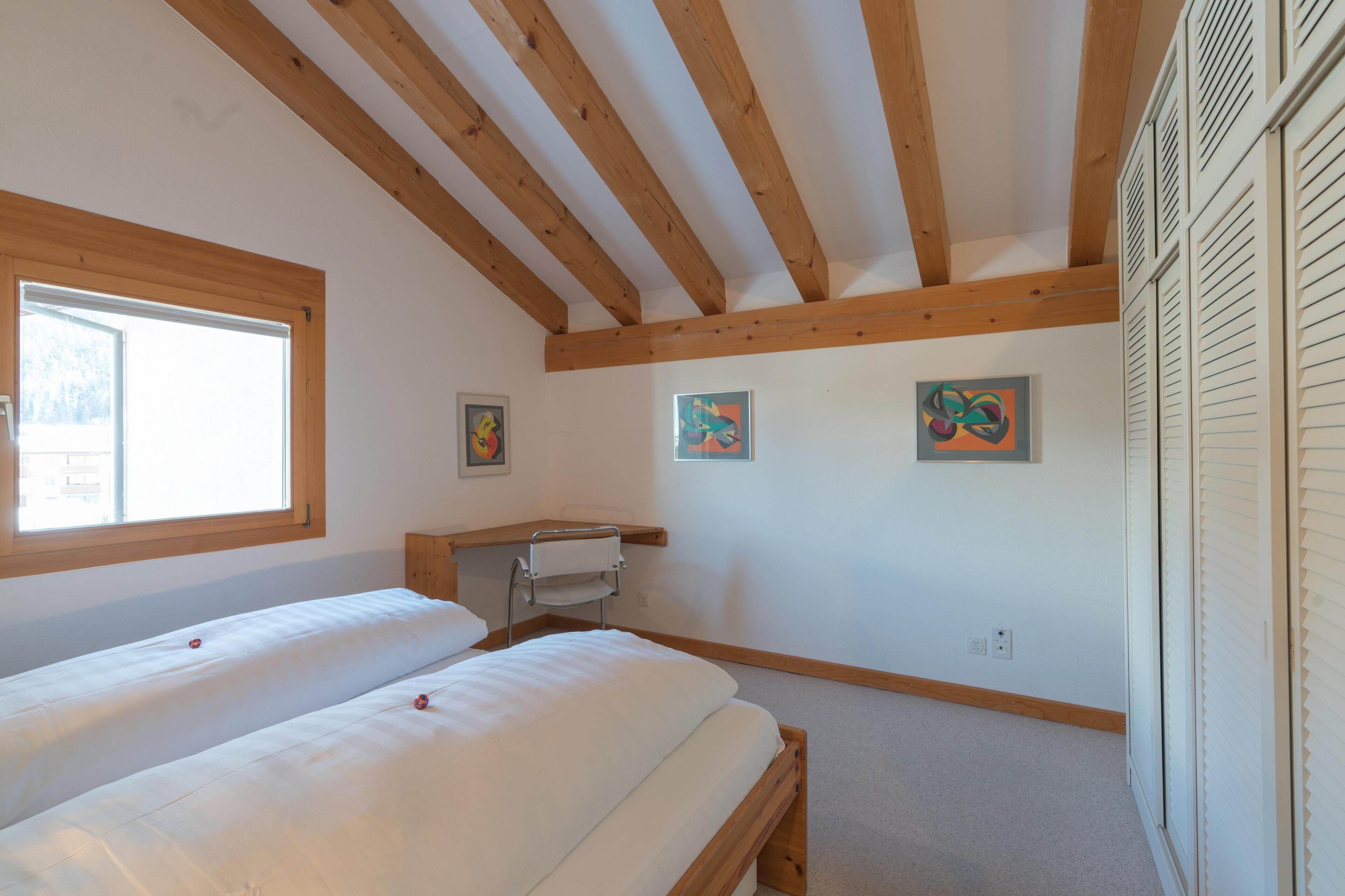 A room with a wooden ceiling, two beds, a window, and artwork on the walls.
