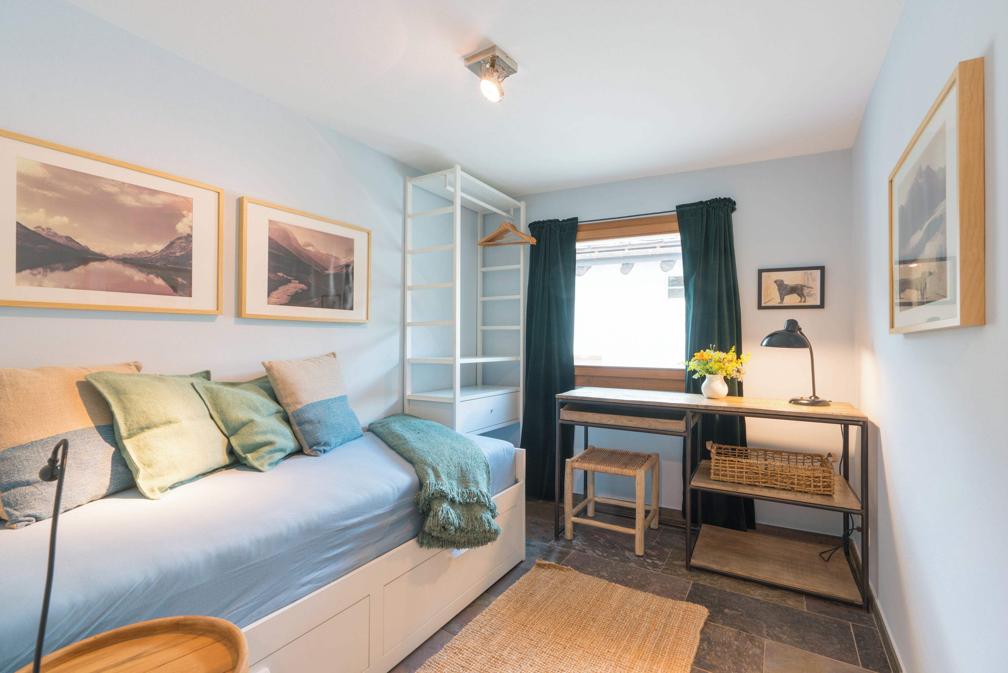 Een slaapkamer met een bed, een bureau, een stoel, een plank en een raam met gordijnen.