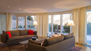 A modern living room with a sofa, a coffee table, and a view of a mountainous landscape through large windows. - Residenza Alpenrose 31 – Spacious 4.5-room flat with a terrace and mountain view (Sils / Segl Maria)