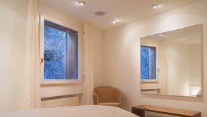A bedroom with a bed, a chair, a bedside table, a window with curtains, and a mirror on the wall. - Residenza Alpenrose 31 – Spacious 4.5-room flat with a terrace and mountain view (Sils / Segl Maria)